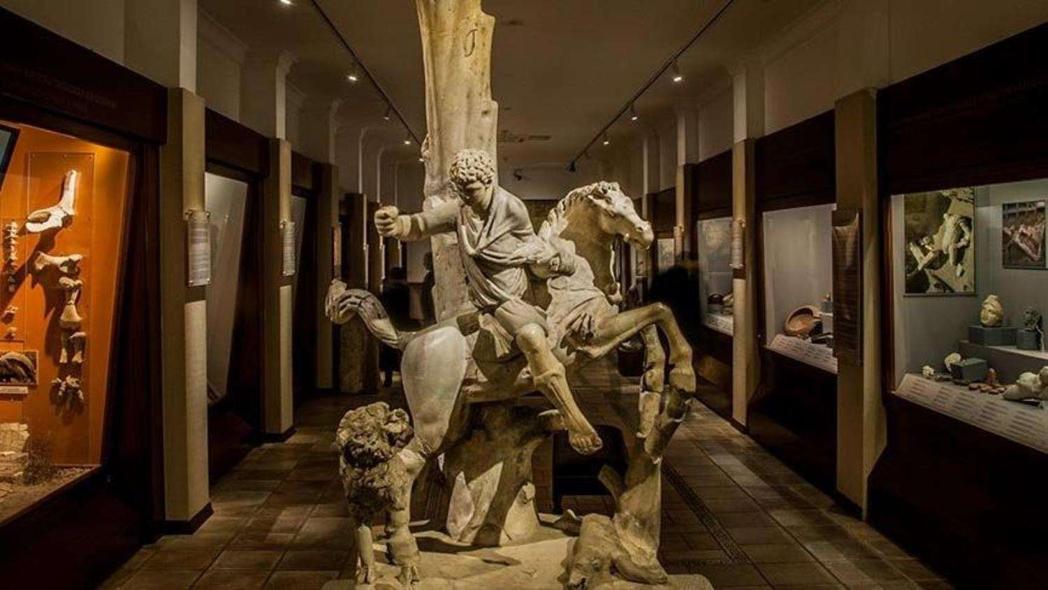 Ancient sculpture of a man on horseback in a museum display, surrounded by various artifacts and exhibits in a dimly lit room.