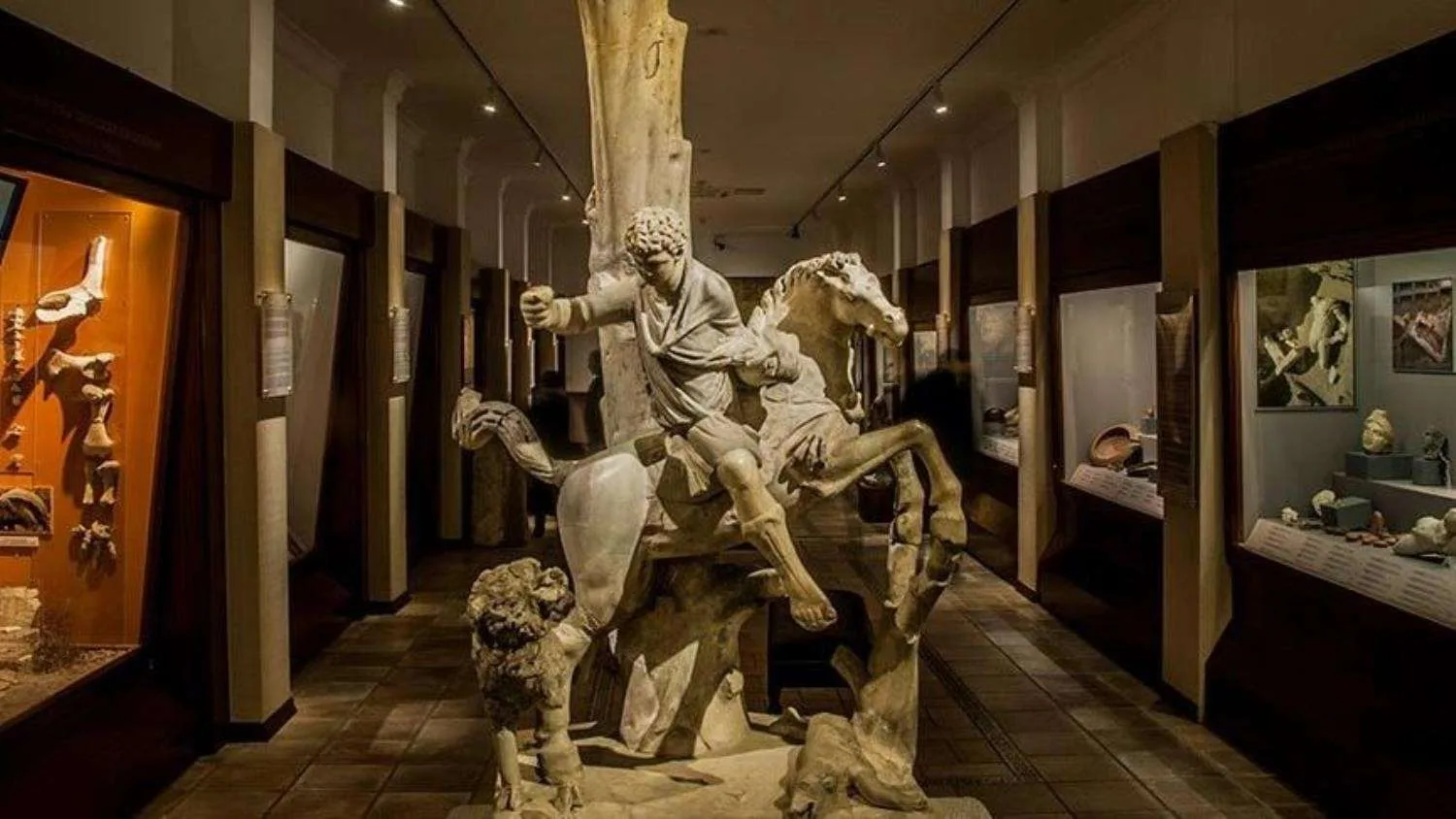 Ancient sculpture of a man on horseback in a museum display, surrounded by various artifacts and exhibits in a dimly lit room.
