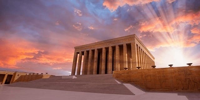A large stone building with tall columns stands under a dramatic sky with sun rays piercing through colorful clouds.