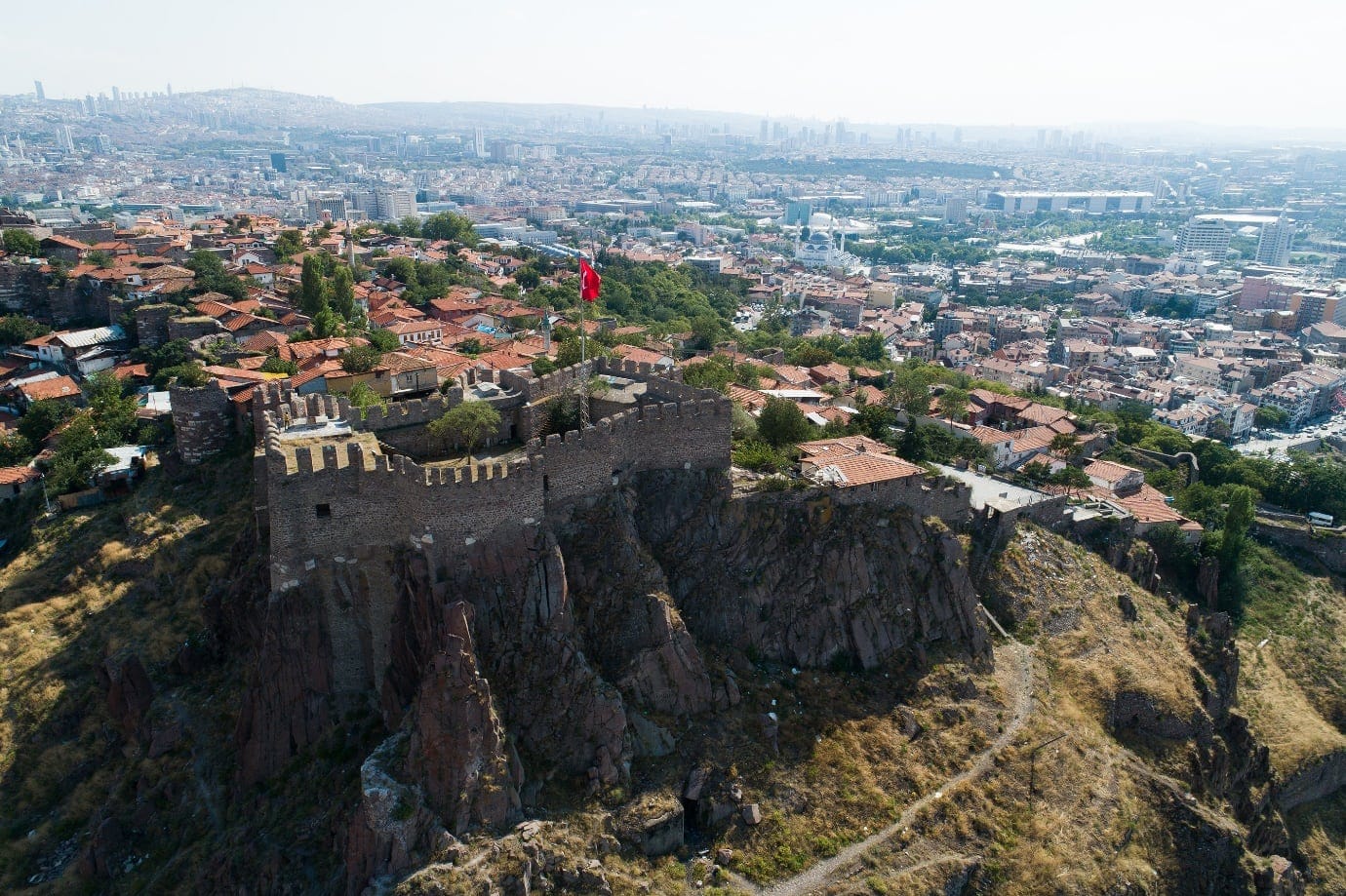 Aerial view of an ancient stone fortress with a Turkish flag, surrounded by a cityscape featuring red-roofed buildings and urban sprawl in the background.