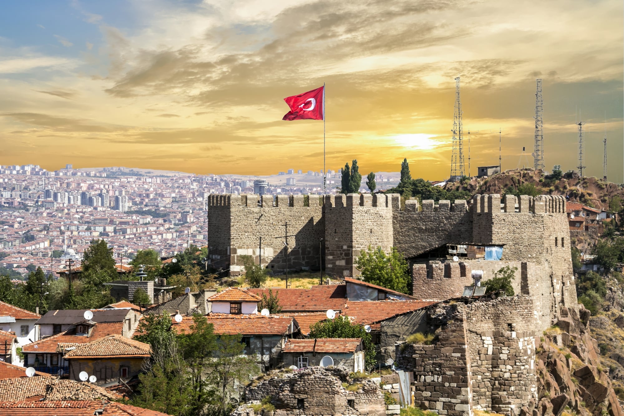 View of a historic fortress in Ankara, Turkey, with a Turkish flag flying atop it. The cityscape and a setting sun are visible in the background.