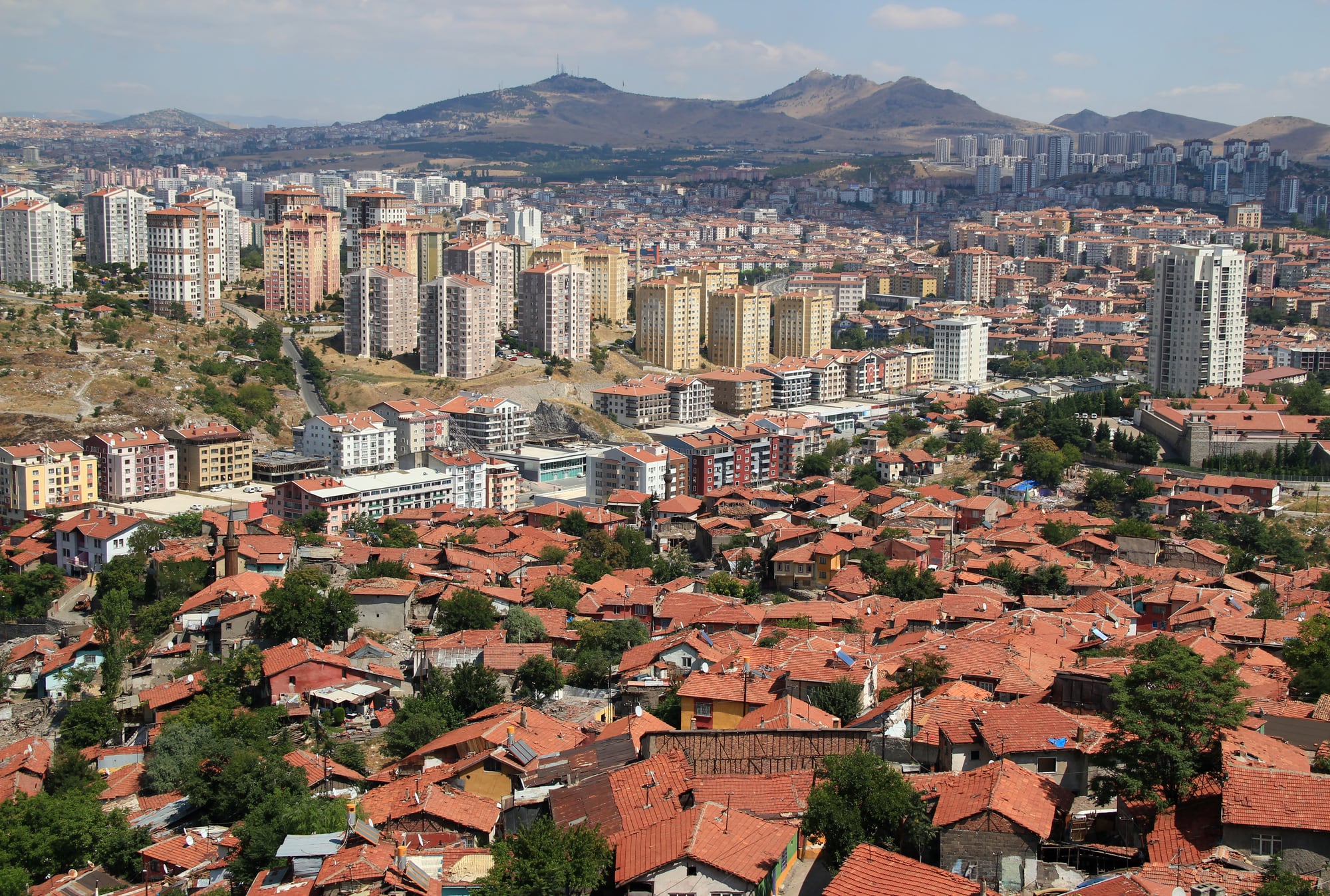 A cityscape showing a mix of densely packed red-roofed houses in the foreground and modern high-rise buildings in the middle and background, set against hilly terrain under a partly cloudy sky.