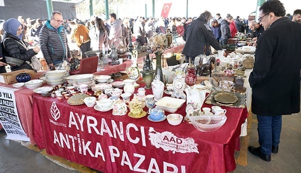 People browsing various antique items on tables at Ayrancı Antique Market, with a red cloth displaying the market's name.