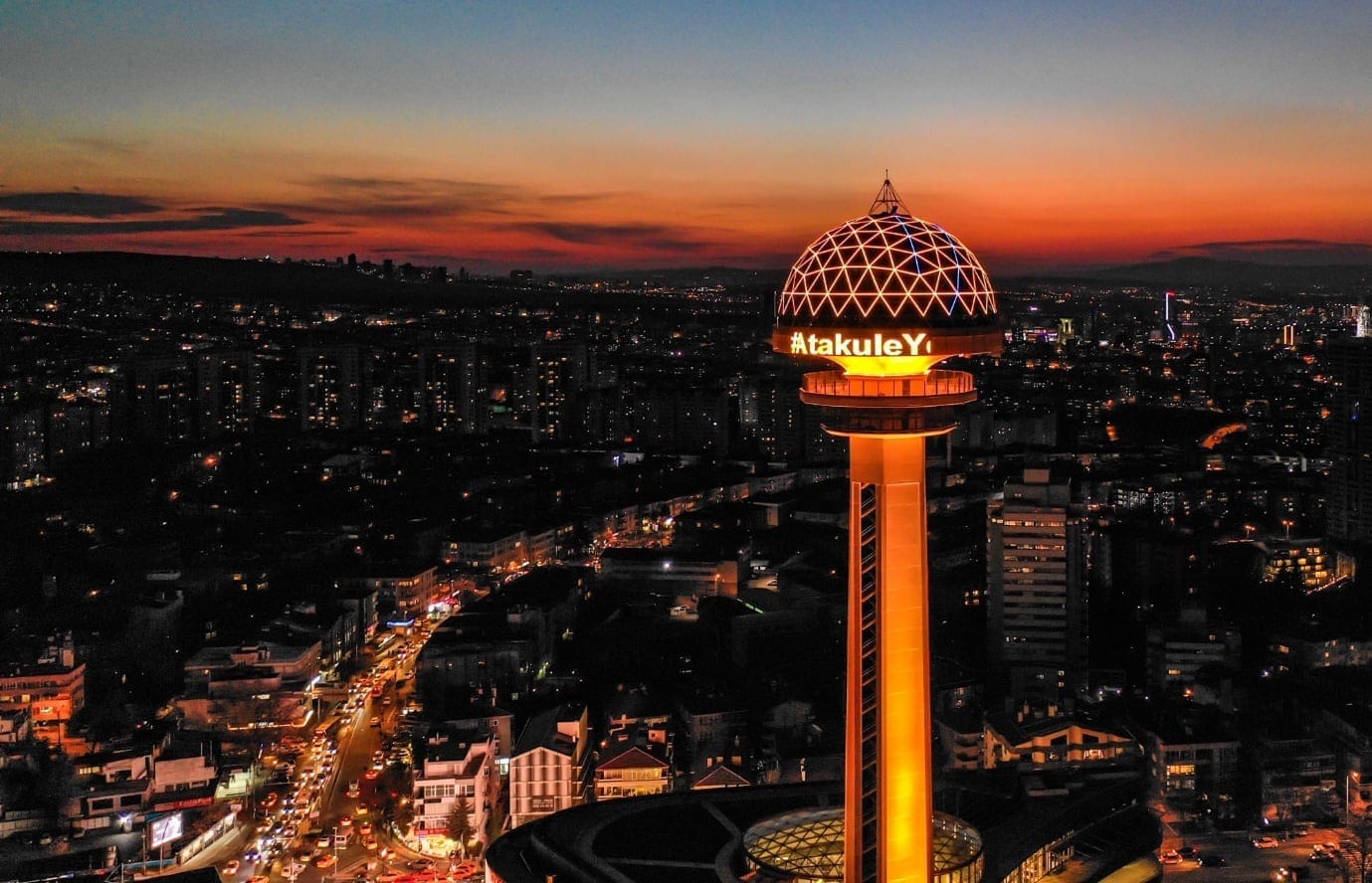 Aerial view of Atakule Tower in Ankara at sunset, with city buildings silhouetted against the colorful sky.