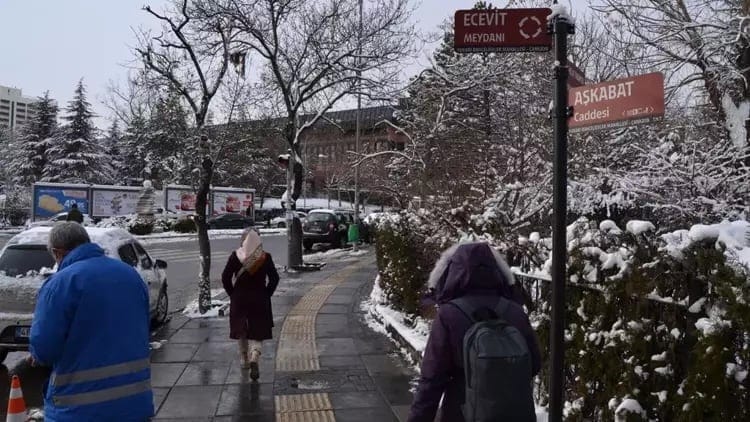 People walking on a snowy sidewalk with trees and road signs in the background.