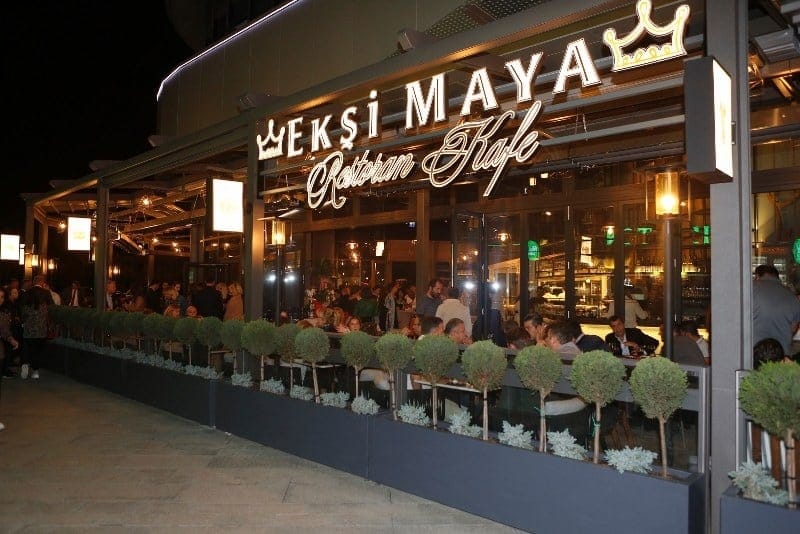 People seated at tables outside Ekşi Maya, a brightly lit café with glass windows and decorative plants, during the evening.