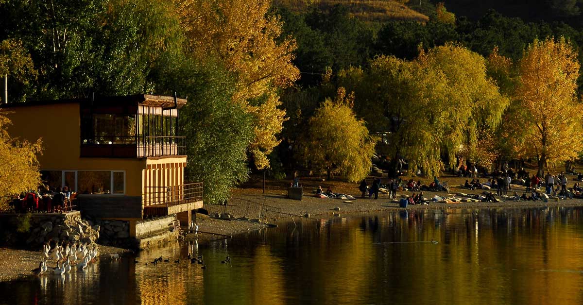 A lakeside scene with a building on the left, autumn trees with golden leaves, and people relaxing on a beach. The water reflects the scenery.